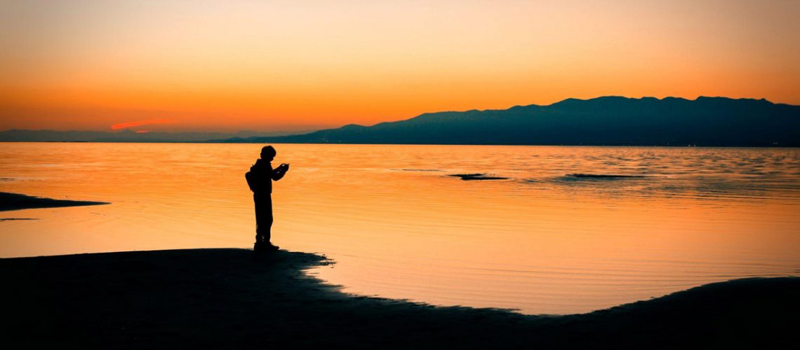 young avoidant man on mobile phone by lake at sunset